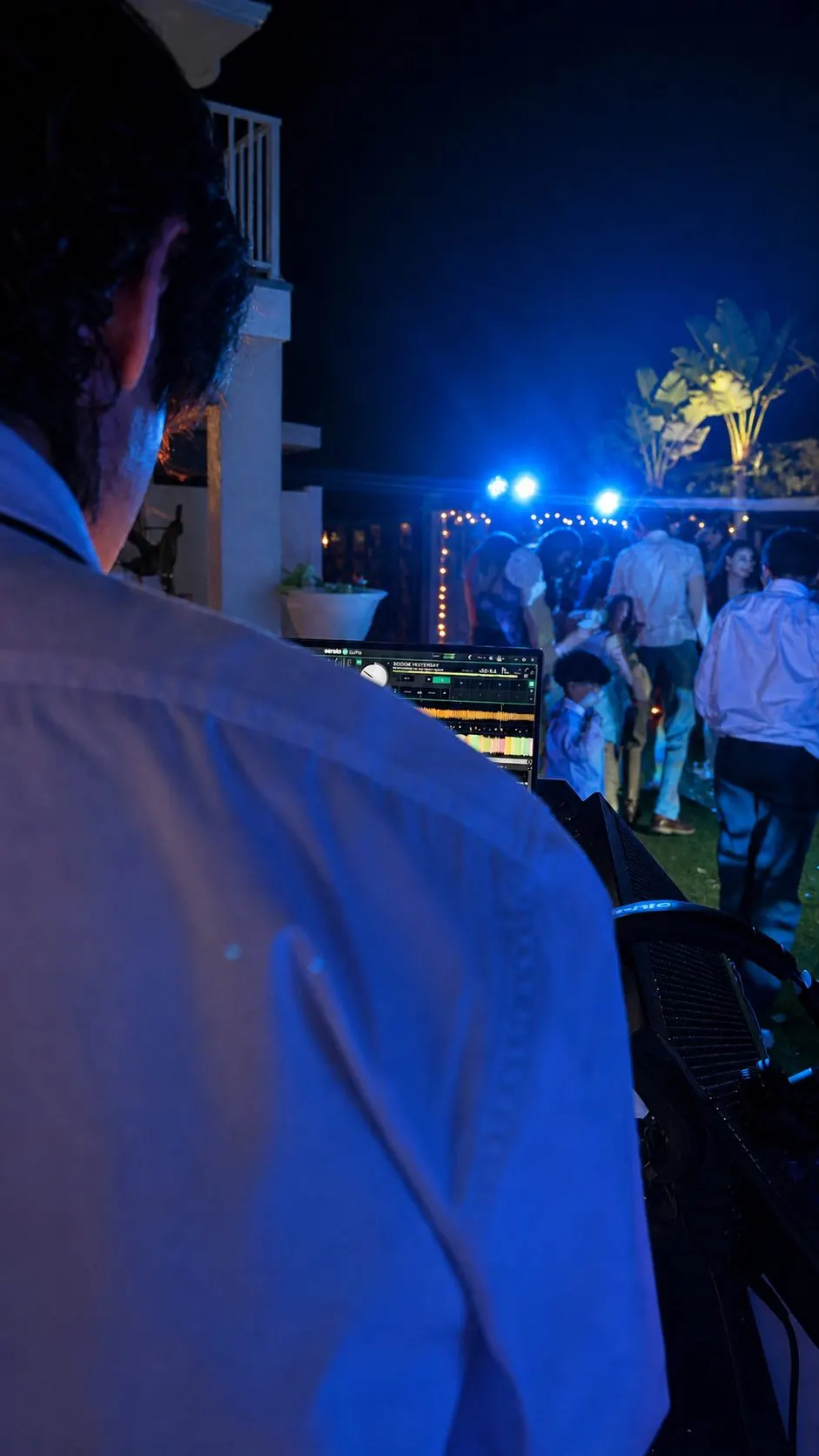 Over a DJ’s shoulder at an outdoor party at night, guests dancing under string lights