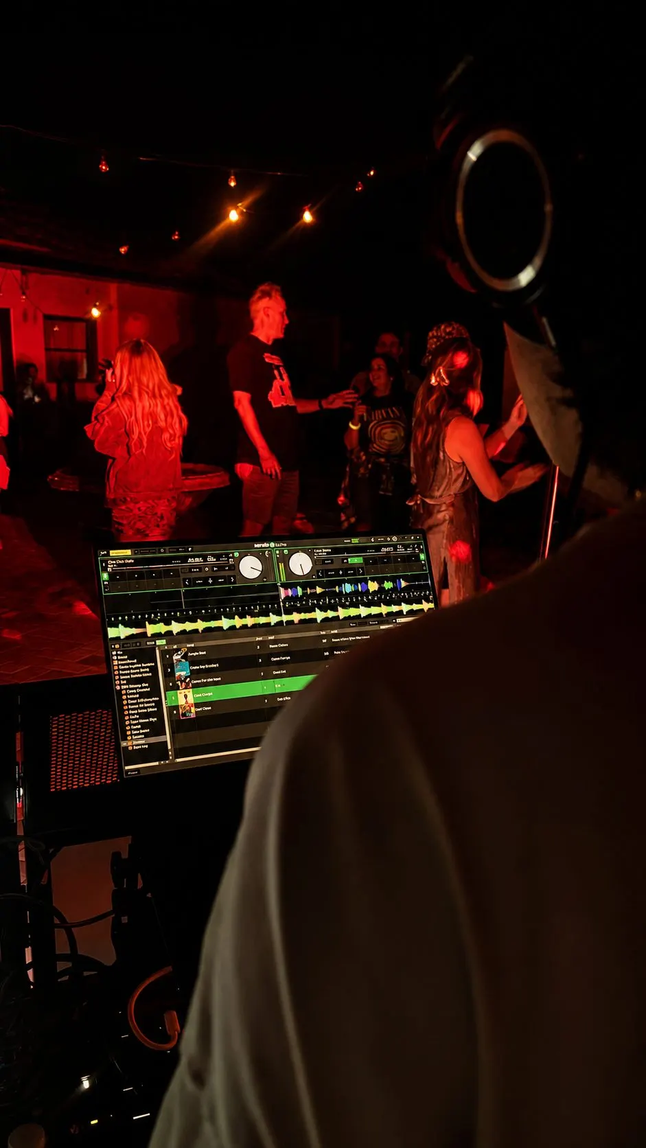 Over a DJ’s shoulder at the laptop, guests and a microphone in the red-lit room beyond
