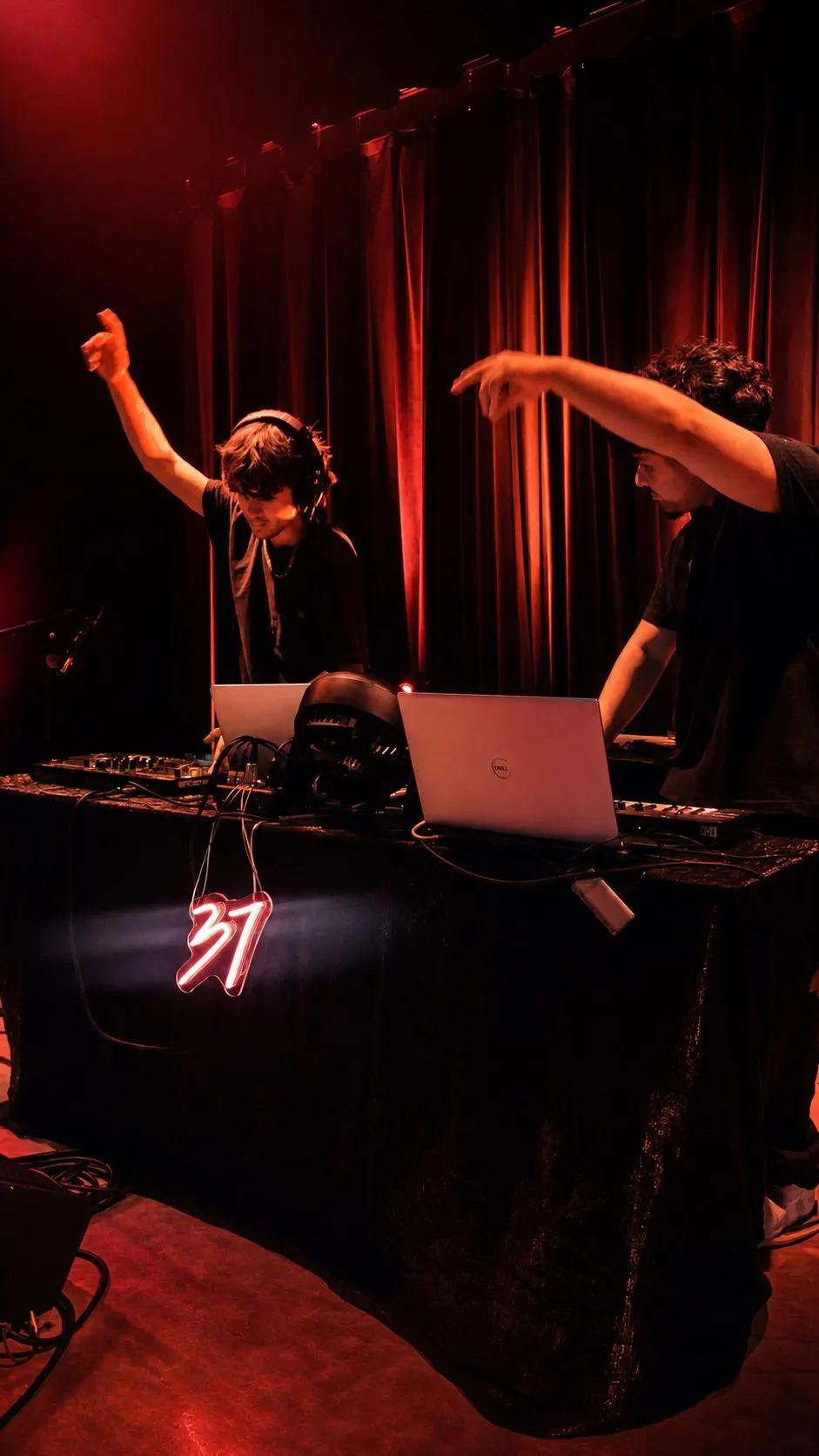 Two DJs working the DJ Studio 37 booth on a stage, red uplighting on the curtain behind them