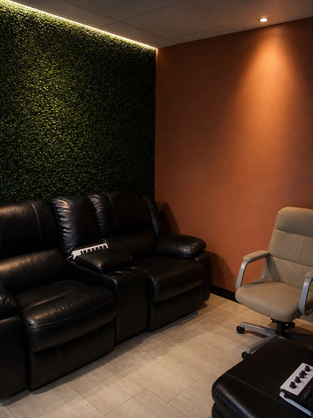 The studio lounge, black leather recliners against a lit green foliage wall